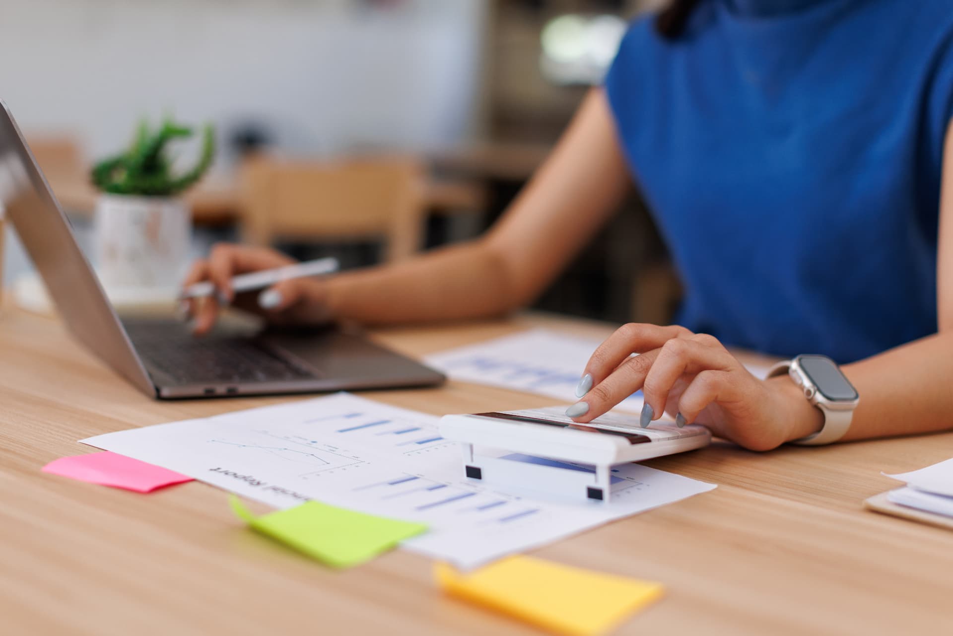 Bookkeeper using a calculator next to a laptop and financial report on a desk